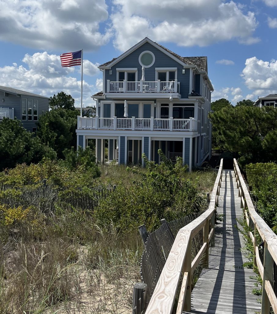 Coughlin House beach home with American flag, surrounded by greenery and a wooden walkway, showcasing coastal architecture and outdoor living spaces.